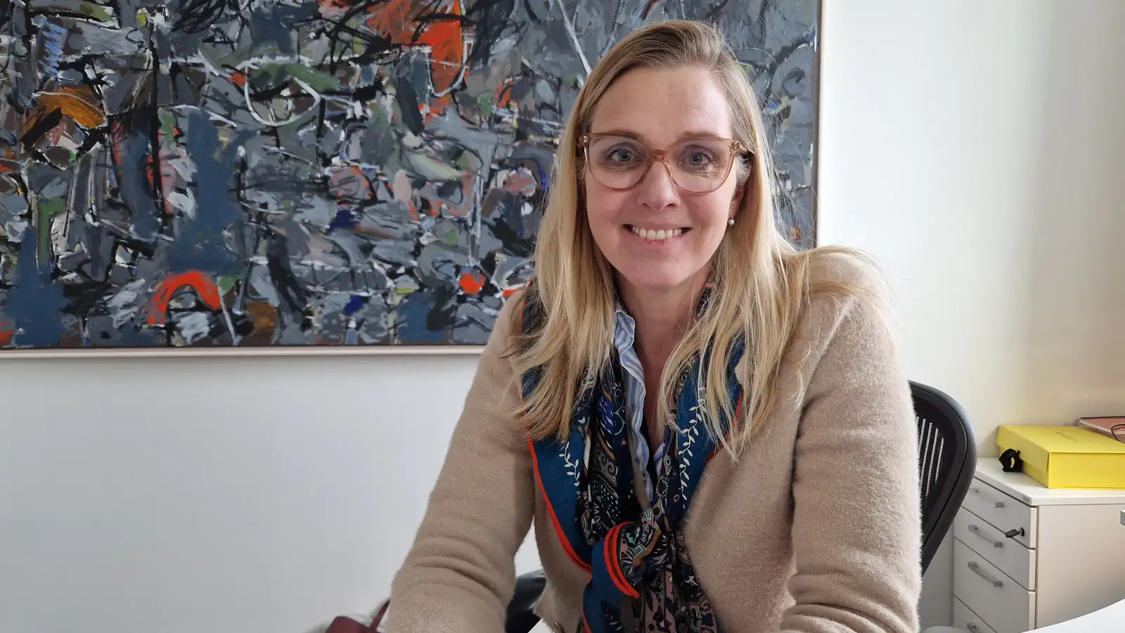 Ásthildur Sturludóttir, bæjarstjóri á Akureyri. Hún situr við skrifborð, í brúnni peysu og með gleraugu. Málverk í baksýn.