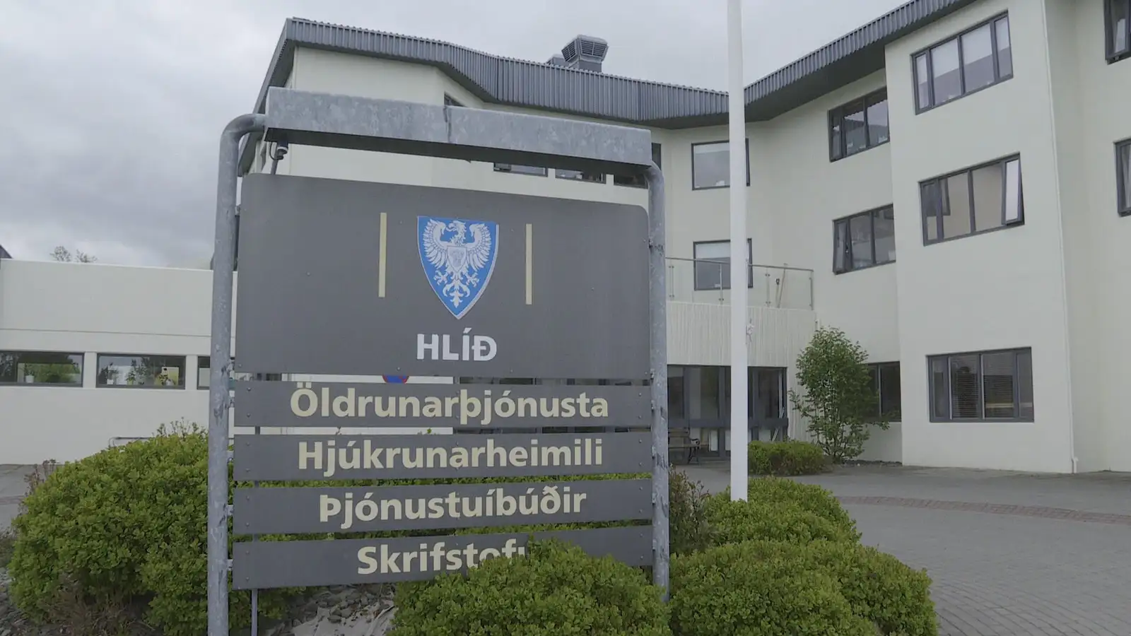 Skilti framan við aðalinngang hjúkrunarheimilisins Hlíðar á Akureyri. Byggingin er beinhvít, þríreist steypubygging með gráu þaki og gluggum.  Skiltið er grátt að lit, efst á því er blátt skjaldarmerki Akureyrar með hvítum erni og nafni heimilisins, Hlíð. Þar fyrir neðan stendur ofan frá: Öldrunarþjónusta, Hjúkrunarheimili, Þjónustuíbúðir, Skrifstofur. Neðan við skiltið eru lágir runnar.