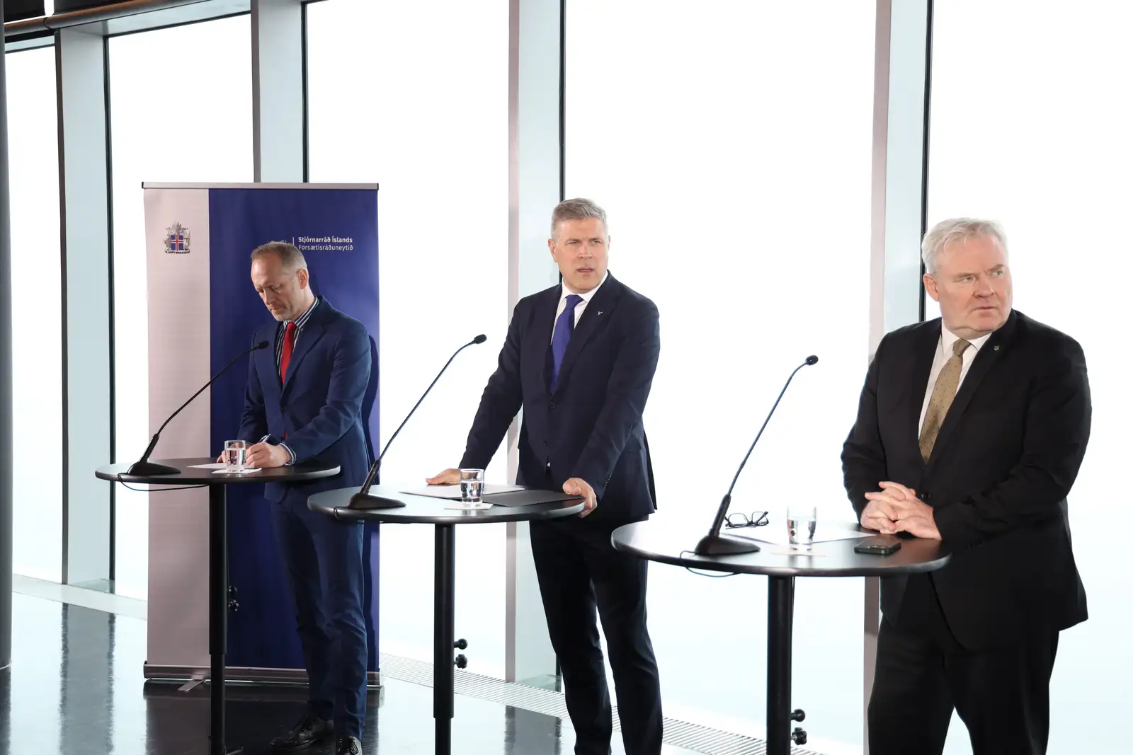 Blaðamannafundur í Hörpu 9. apríl. Formenn stjórnarflokkanna þriggja standa við lítil borð framan við glerveggi Hörpu; lengst til vinstri er Guðmundur Ingi Guðbrandsson formaður Vinstri grænna, í miðið er Bjarni Benediktsson formaður Sjálfstæðisflokks og til hægri er Sigurður Ingi Jóhannsson formaður Framsóknarflokksins.