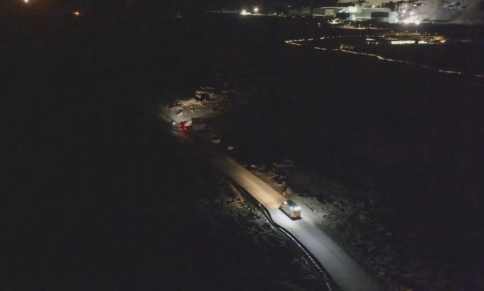 Am Abend des 23. November 2024 fahren Feuerwehrfahrzeuge in stockfinsterer Dunkelheit über die Straße entlang der Verteidigungsmauer bei Svartsengi. Im Hintergrund ist das Kraftwerk in Svartsengi zu sehen.