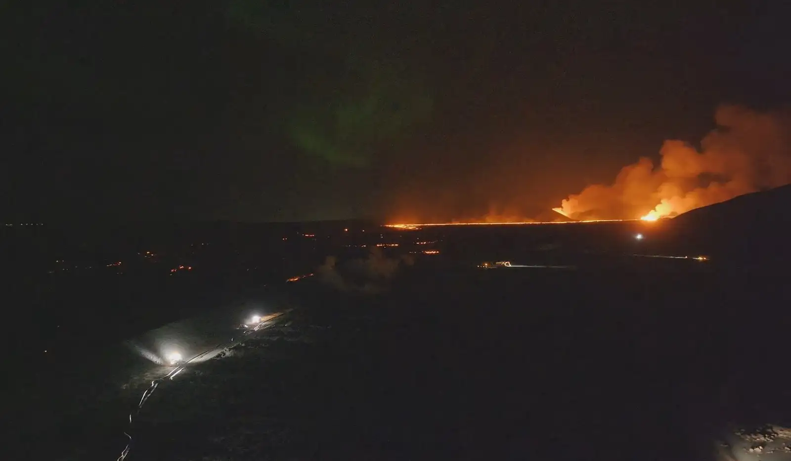 Gesehen über den Befestigungsanlagen von Svartsengi in stockfinsterer Dunkelheit am Abend des 23. November 2023. In der Ferne können Sie das Glitzern von brennender Lava und Vulkanasche aus der Eruptionsserie Sundhnúksgíga sehen, und im Vordergrund können Sie die Lichter sehen von Autos und Arbeitsmaschinen auf der Festung.